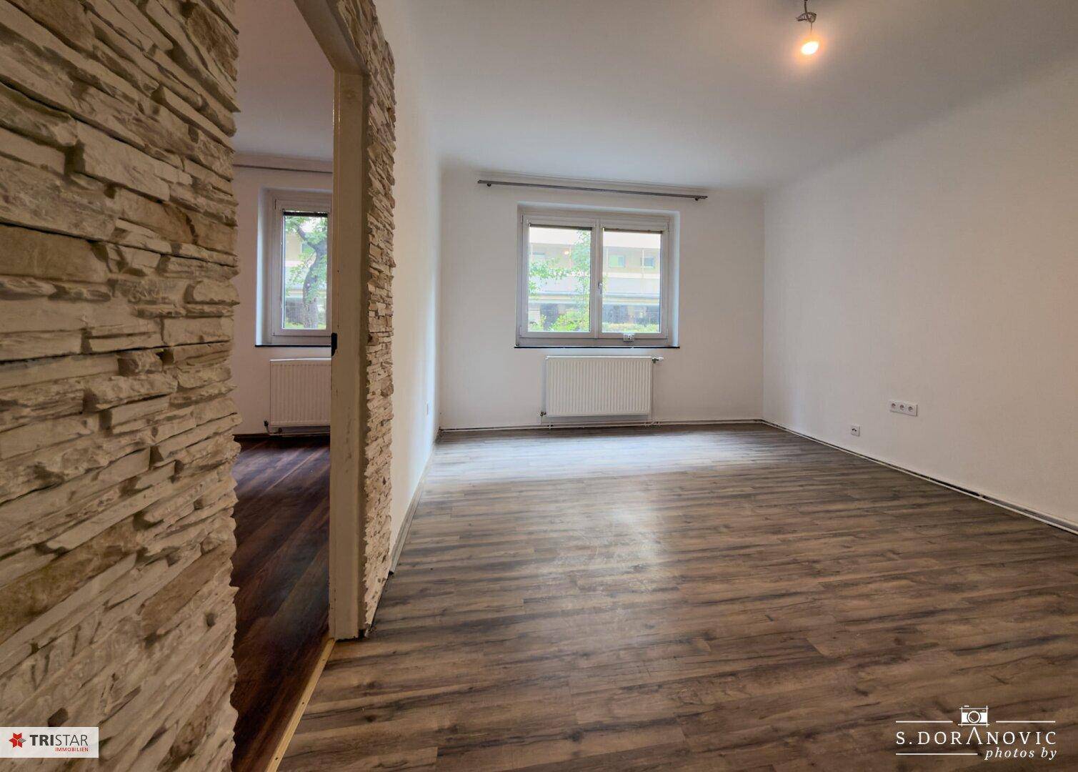 Geräumiges Zimmer mit Laminatboden, zwei Fenstern und dekorativer Steinwand, die einen Durchgang betont.