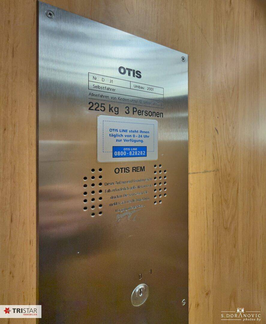 Informationsschild des Aufzugs mit Herstellerangaben und Notrufnummern, an einer Holzwand befestigt.