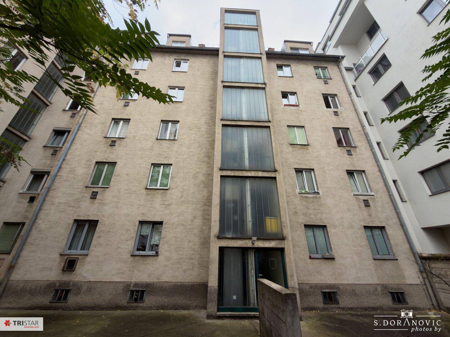 Fassade eines Mehrfamilienhauses mit einem außenliegenden Glasaufzug, der das Gebäude modernisiert.