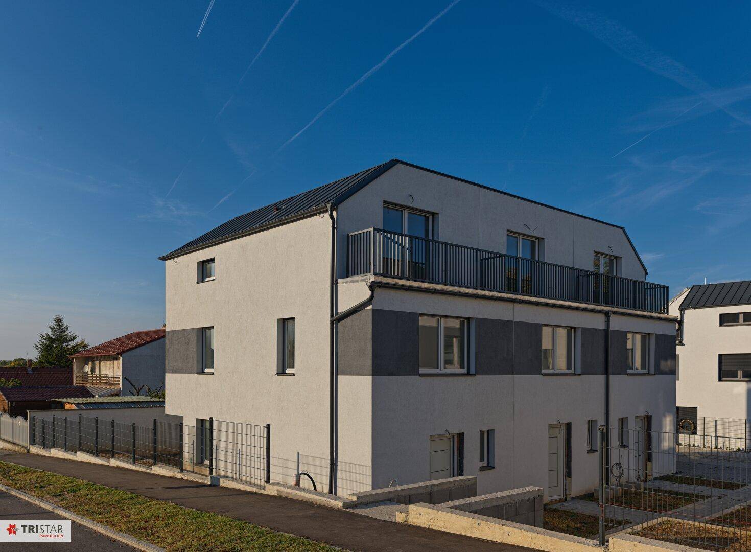Seitenansicht eines modernen Reihenhauses mit Balkon im Obergeschoss und Zaun.