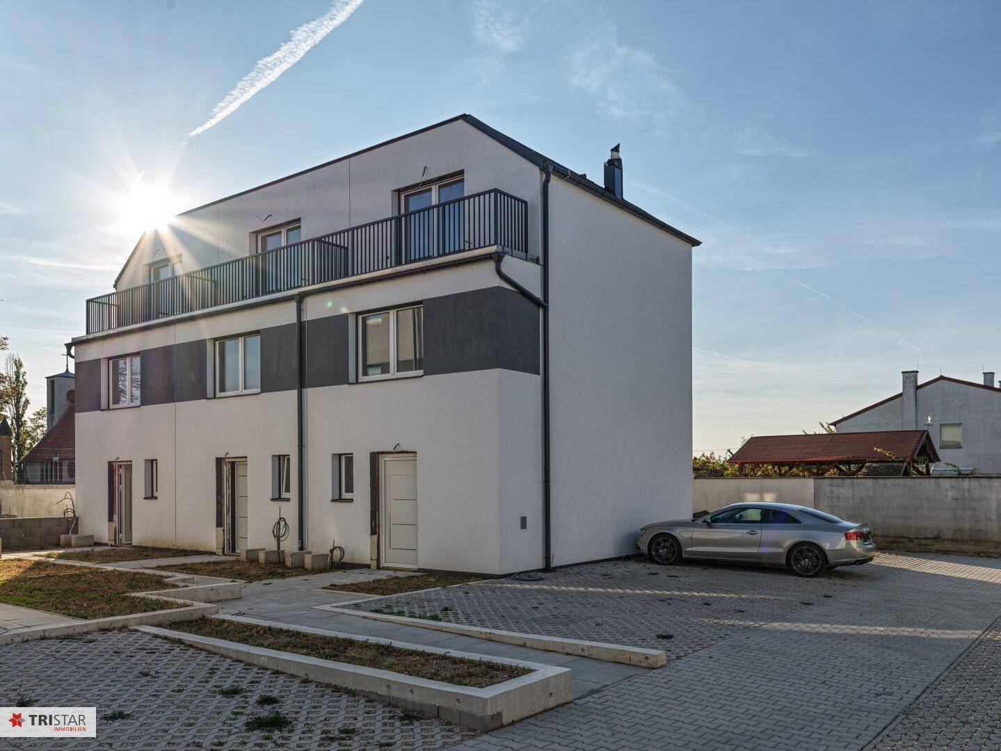 Modernes Reihenhaus mit Balkon im Obergeschoss und Parkplatz unter strahlendem Sonnenschein.