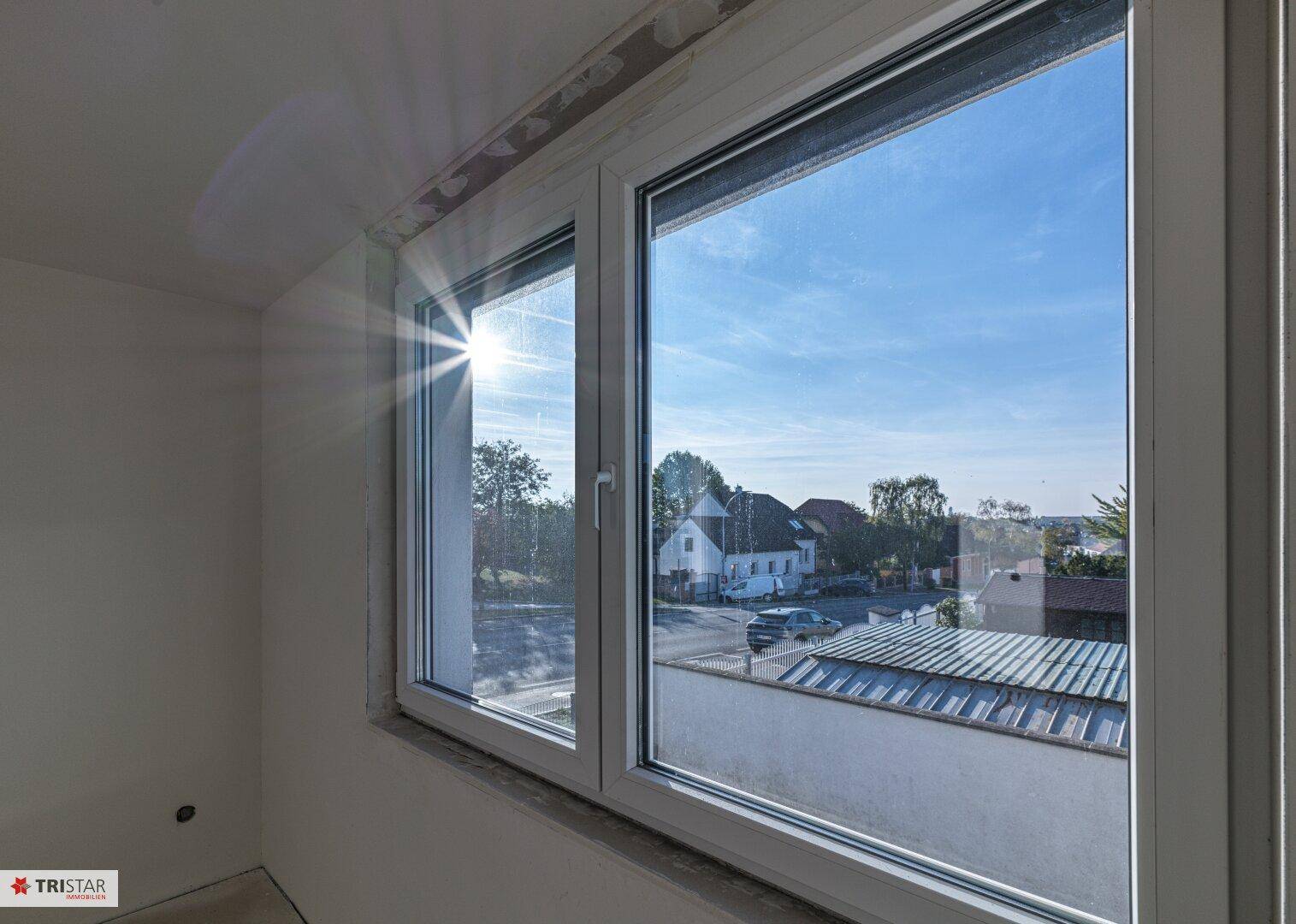 Blick aus dem Fenster auf die Umgebung mit Häusern und Bäumen unter blauem Himmel.