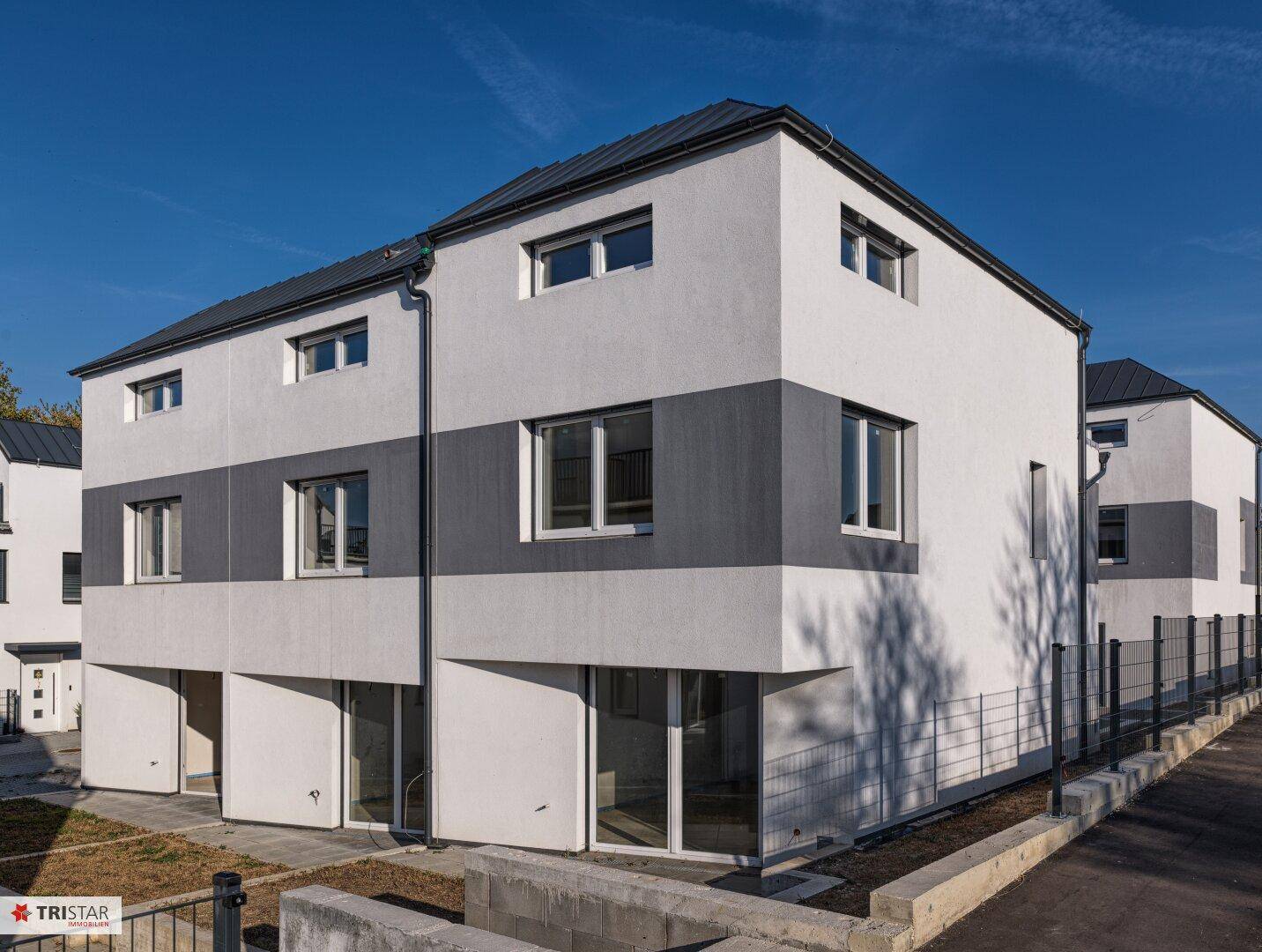 Vorderansicht eines modernen Reihenhauses mit weiß-grauer Fassade und großen Fenstern unter blauem Himmel.