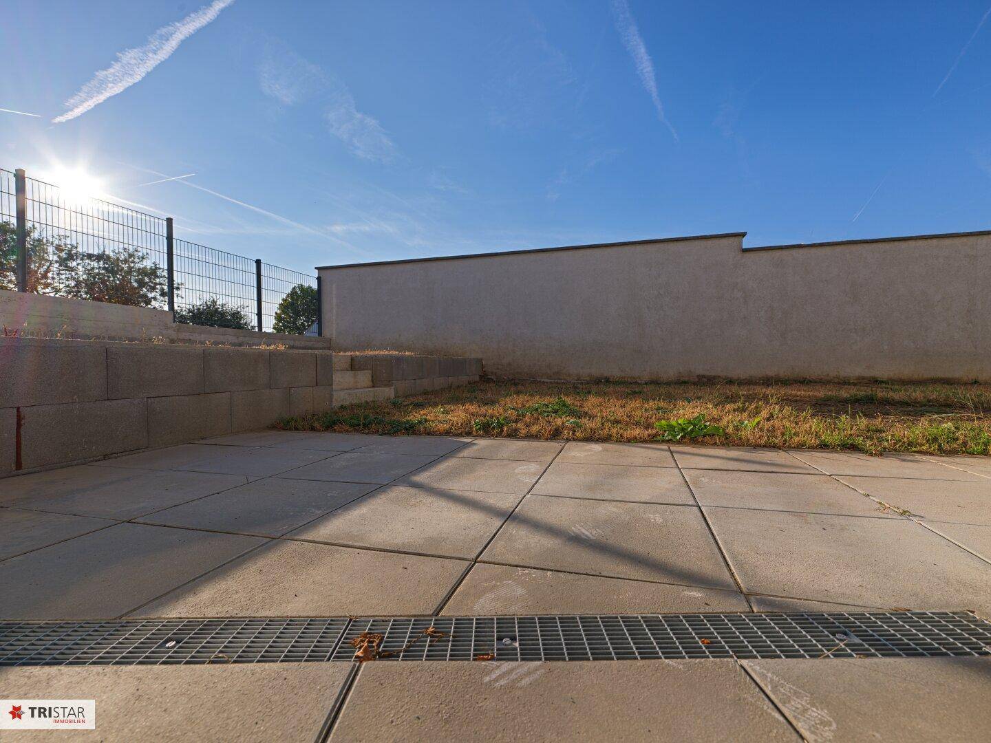 Gepflasterte Terrasse mit Entwässerungsrinne und angrenzender Betonmauer bei Sonnenschein.