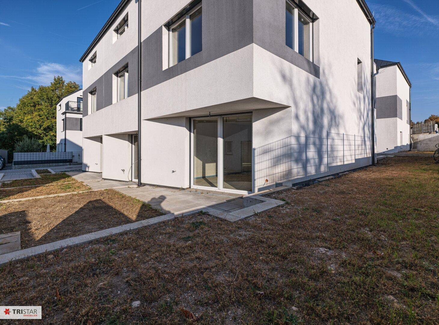 Modernes Reihenhaus mit weiß-grauer Fassade und ungestaltetem Gartenbereich unter blauem Himmel.