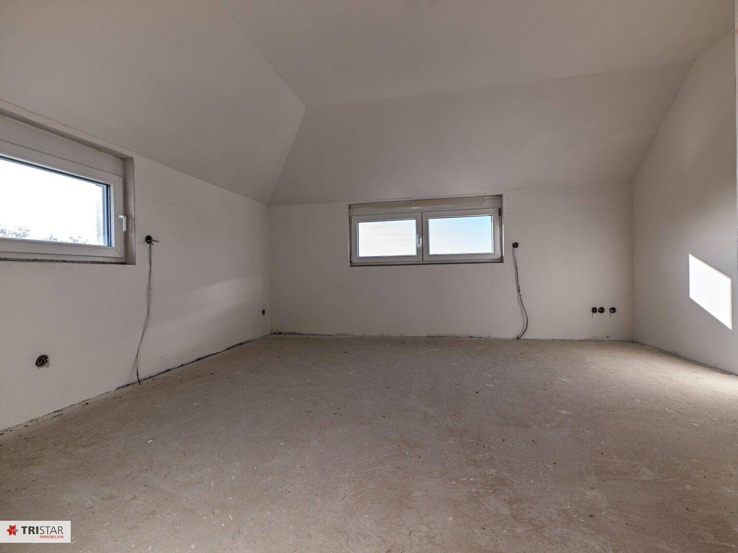 Geräumiger Dachgeschossraum im Rohbauzustand mit schräger Decke und zwei Fenstern.