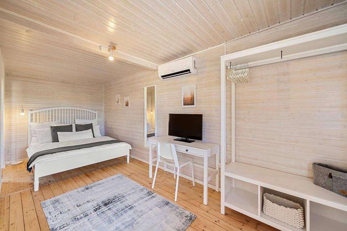 Gemütliches Schlafzimmer mit weißem Bett, Schreibtisch, Fernseher und Klimaanlage in heller Holzoptik.