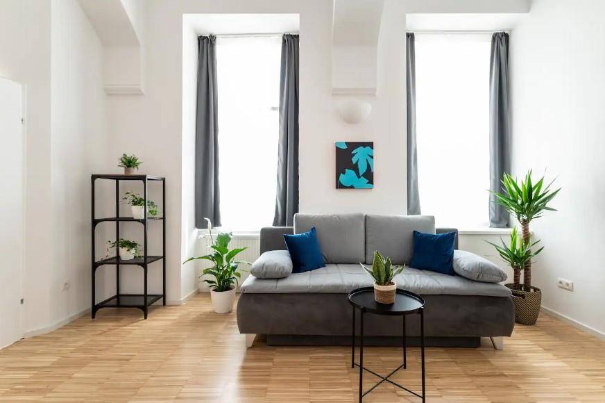 Gemütliches Wohnzimmer mit grauem Sofa, Pflanzen und hohen Fenstern, die den Raum erhellen.