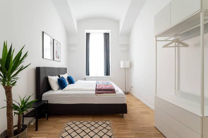 Schlafzimmer mit Doppelbett, offener Garderobe und dekorativer Pflanze, minimalistisch gestaltet.