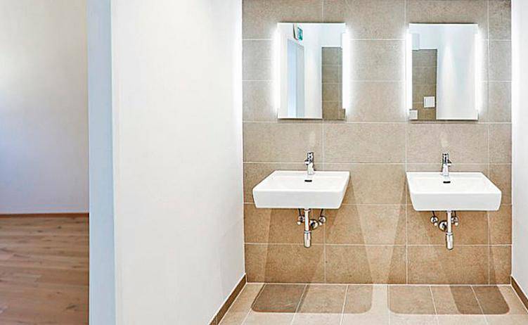 Moderner Waschraum mit zwei eleganten Waschbecken und beleuchteten Spiegeln, gefliest in neutralen Tönen.