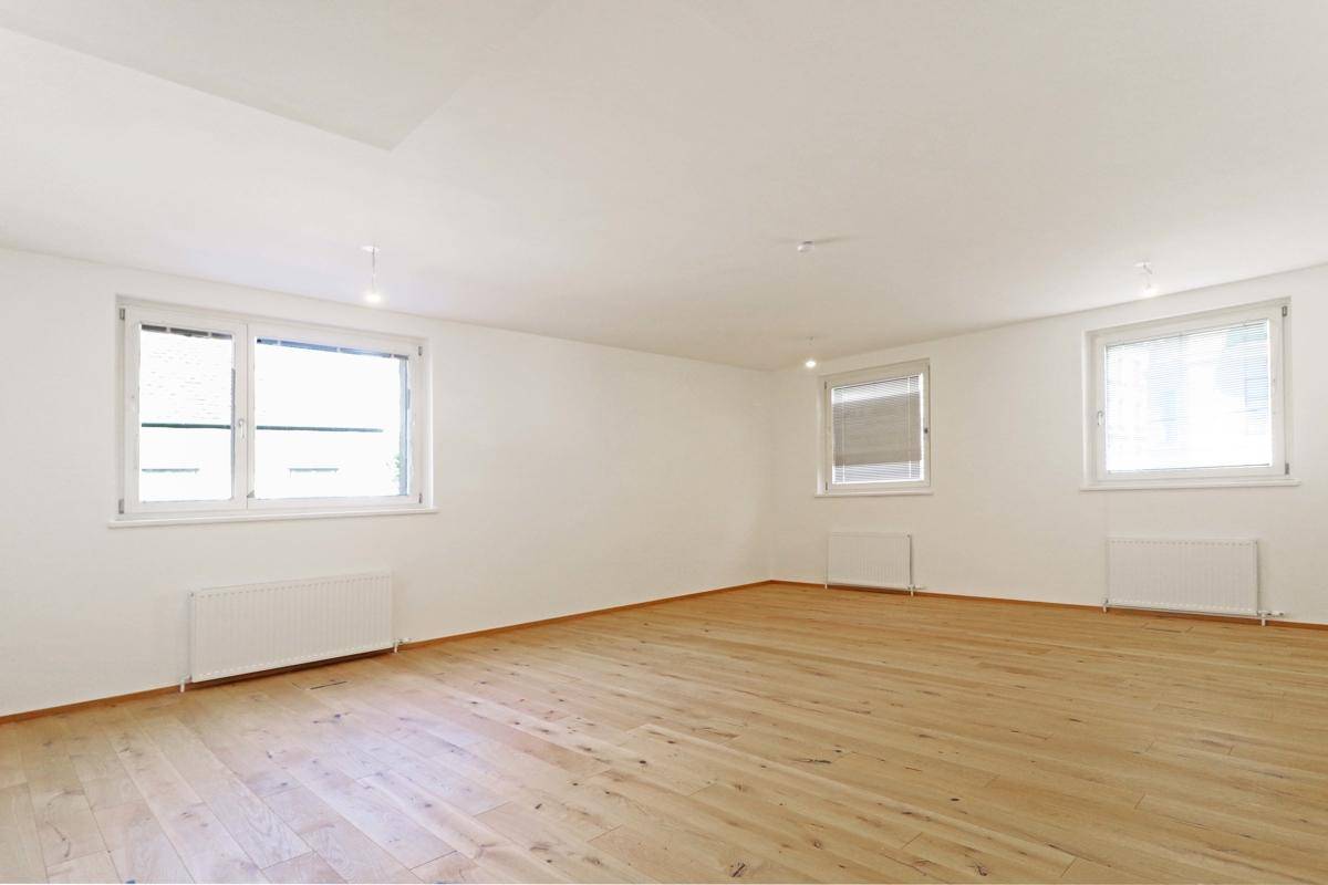 Heller Raum mit Holzboden und zwei großen Fenstern, ausgestattet mit Heizkörpern unter den Fensterbänken.