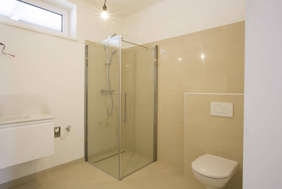Modernes Badezimmer mit begehbarer Glasdusche, wandhängendem WC und beigefarbenen Fliesen an Wänden und Boden.