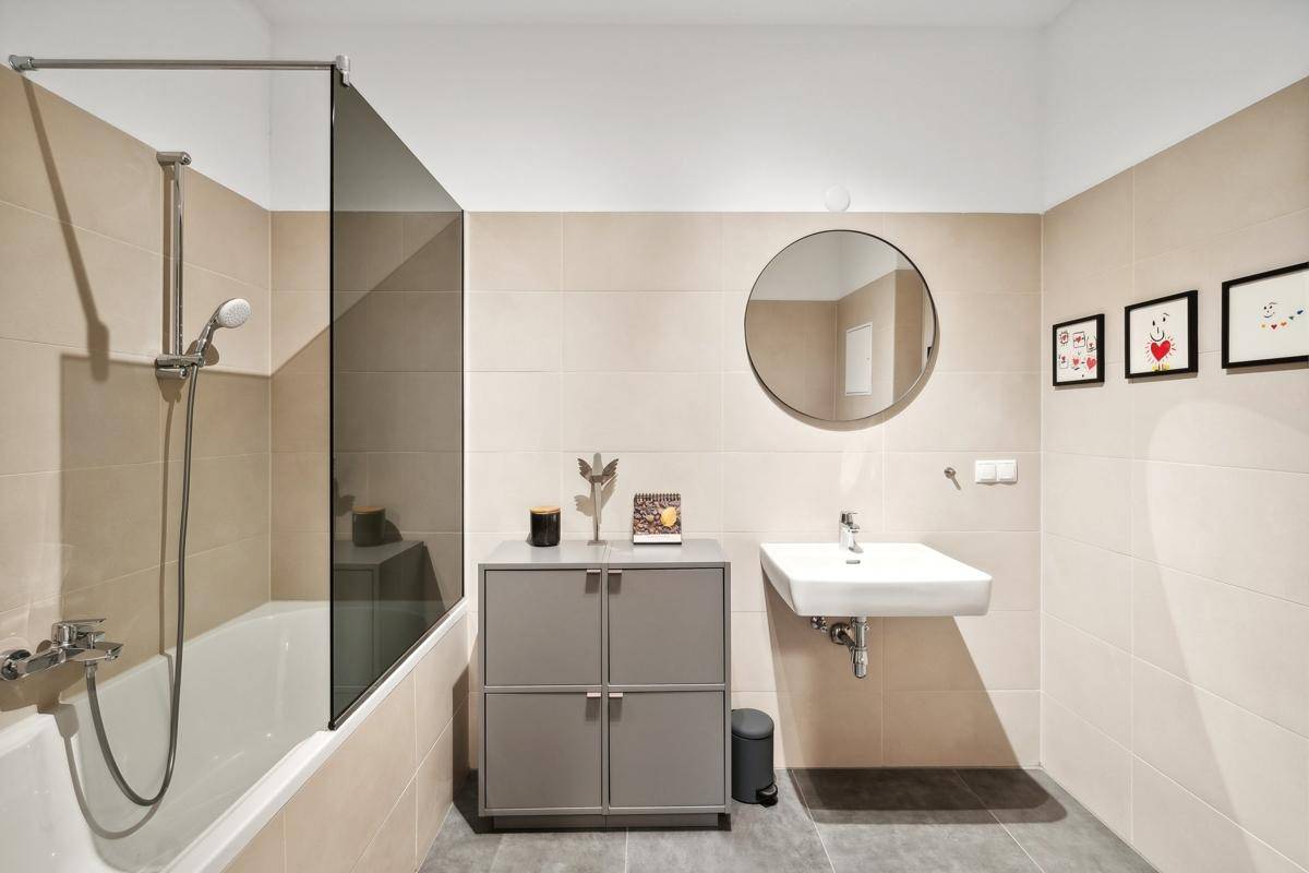 Modernes Badezimmer mit beigefarbenen Fliesen, Badewanne mit Duschabtrennung und rundem Spiegel, sehr gepflegt.