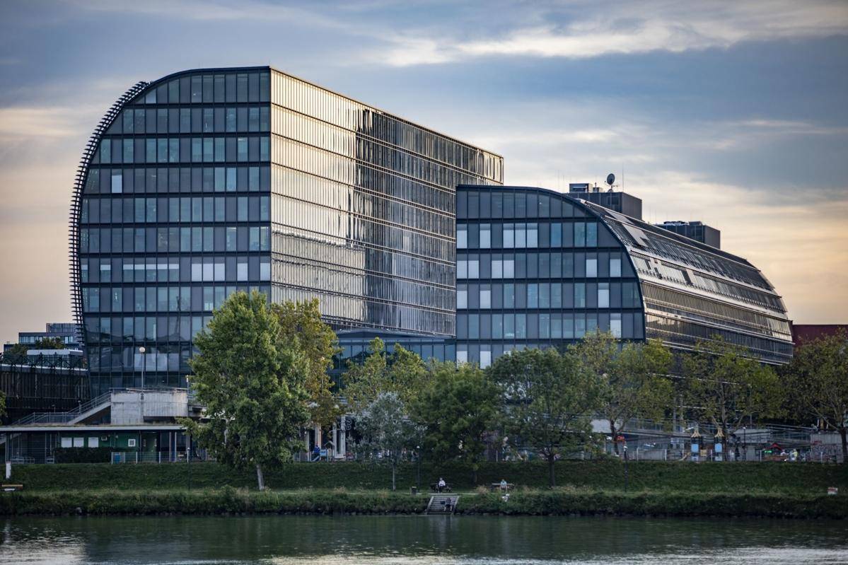 Moderne Bürogebäude mit geschwungener Glasfassade am Flussufer, umgeben von Bäumen und Grünflächen.