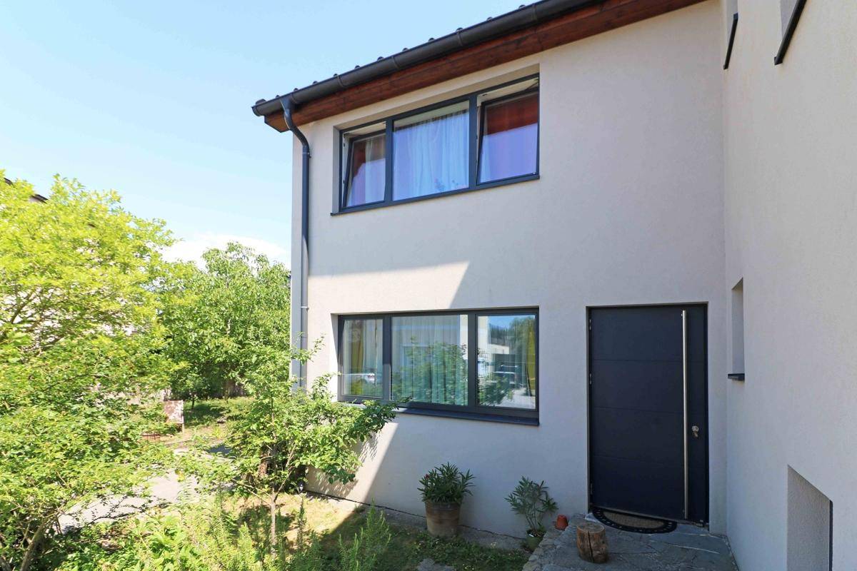 Eingangsbereich des Hauses mit einer modernen dunklen Tür und großen Fenstern, umgeben von Grün.