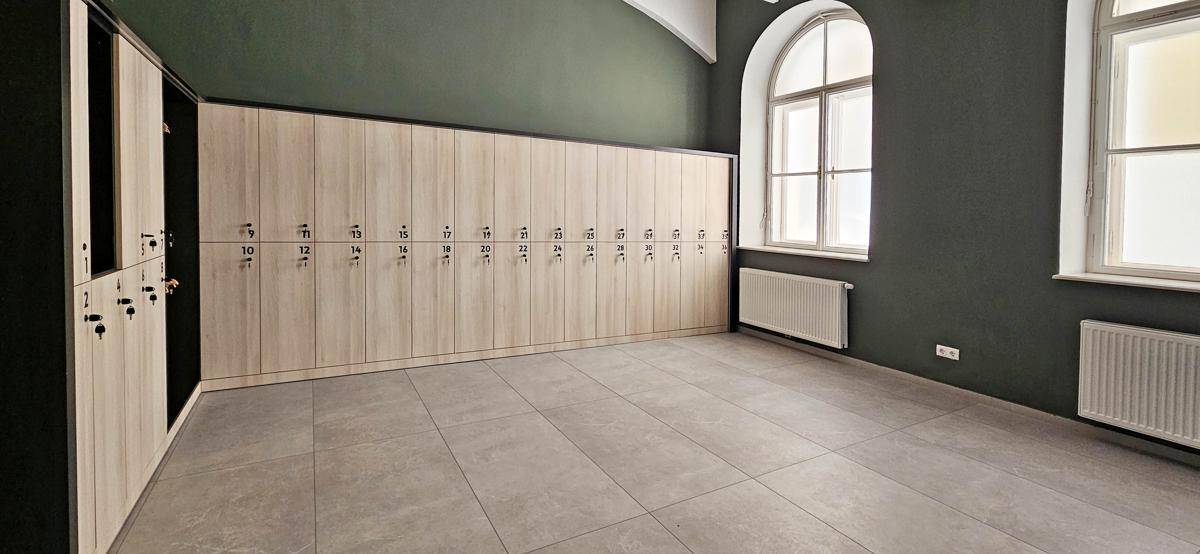Geräumiger Umkleideraum mit vielen Holzschließfächern, hohen Fenstern und Fliesenboden, gut beleuchtet.