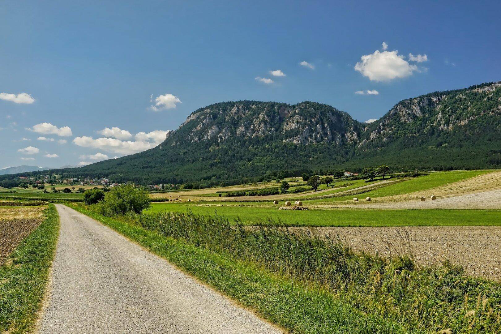 Eine ländliche Straße führt durch weite Felder mit Heuballen und einer beeindruckenden Bergkette am Horizont.