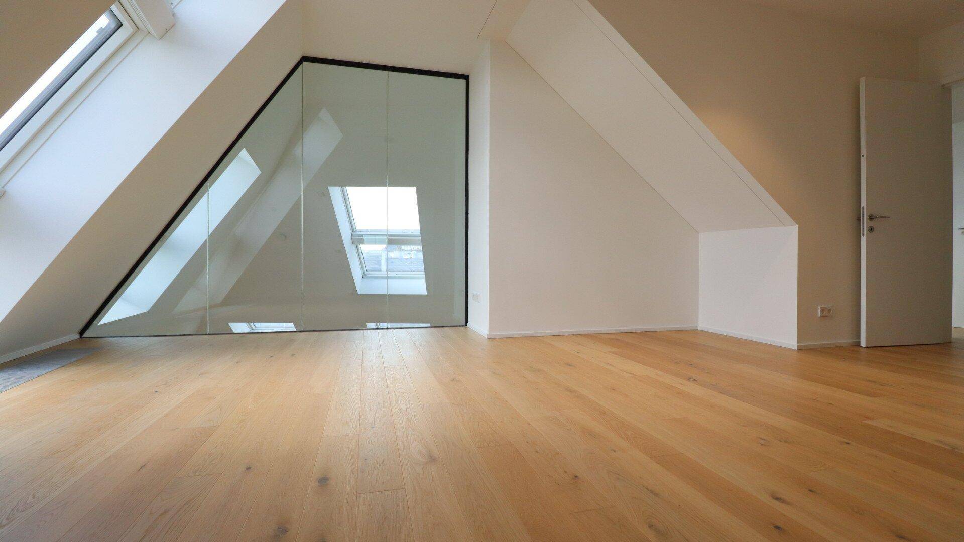 Geräumiges Zimmer im Dachgeschoss mit Glaswand, Dachfenstern und hellem Holzparkett.