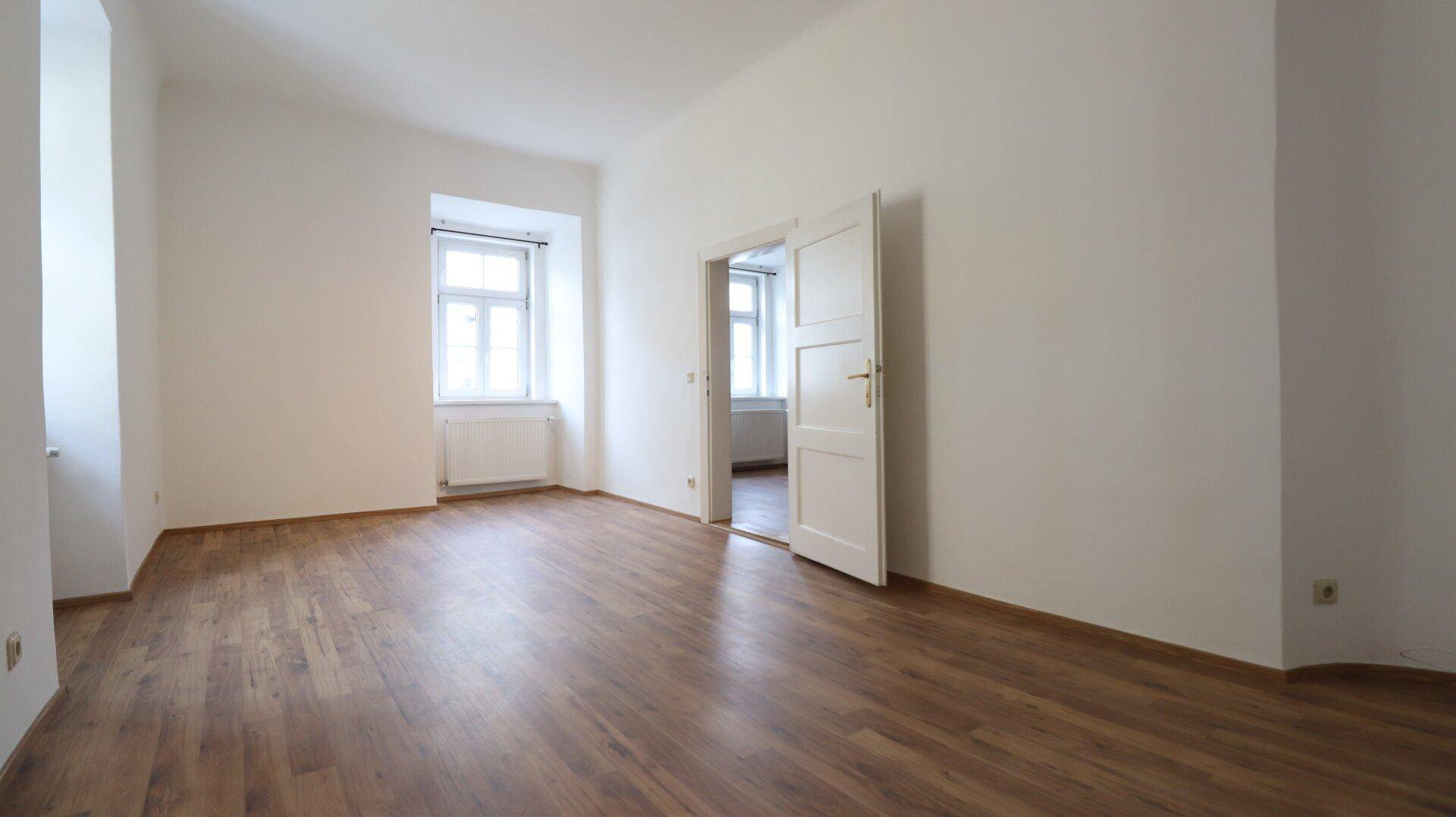 Heller Wohnraum mit Holzboden und zwei Fenstern, die für natürliche Belichtung sorgen.
