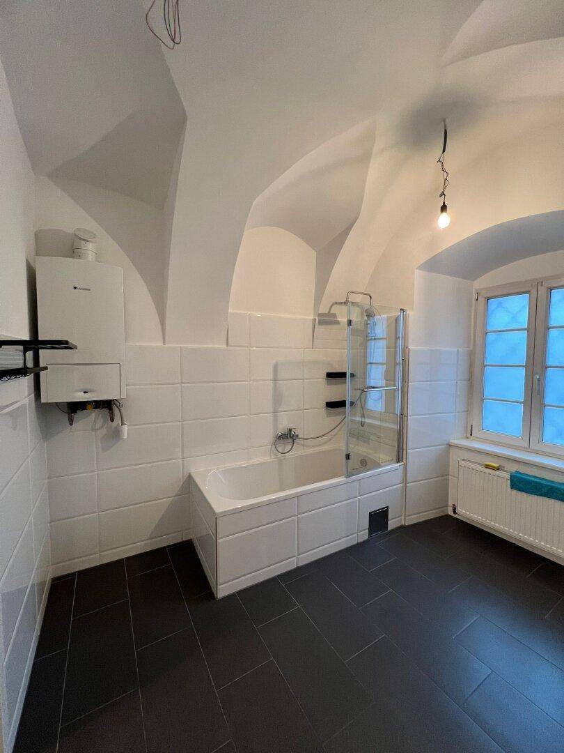 Badezimmer mit Badewanne, weißen Fliesen, dunklem Boden und einer markanten Gewölbedecke.