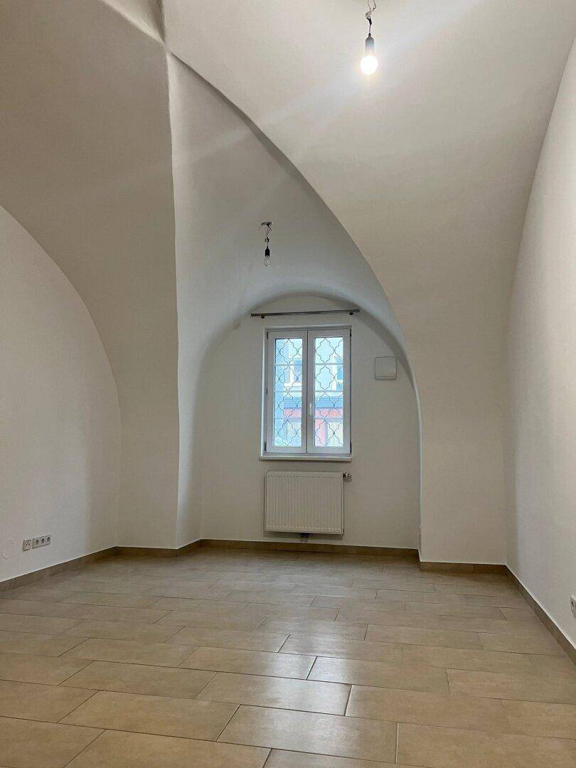 Heller Raum mit einzigartiger Gewölbedecke, Fliesenboden und einem Fenster mit Gitter.