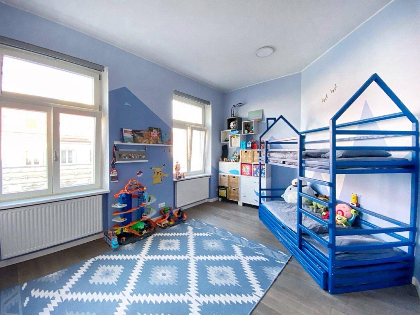 Helles Kinderzimmer mit blauen Etagenbetten, farbigen Wänden und großem Fenster.