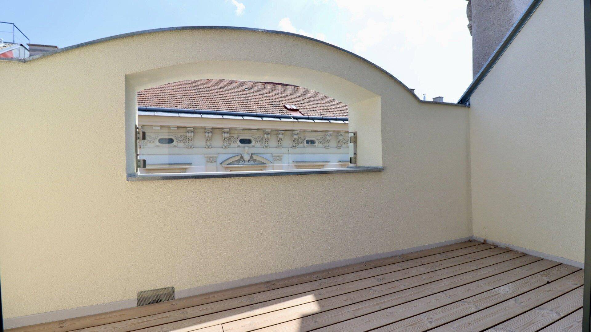 Kleiner, überdachter Balkon mit Holzboden und Blick auf die historische Fassade eines Nachbargebäudes.