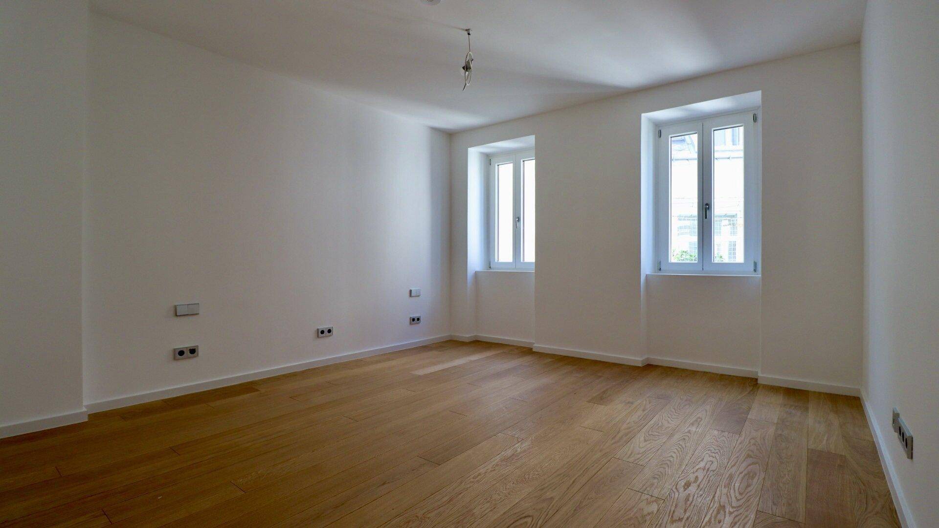 Geräumiger, heller Raum mit Holzboden und zwei hohen Fenstern, die viel Tageslicht hereinlassen.
