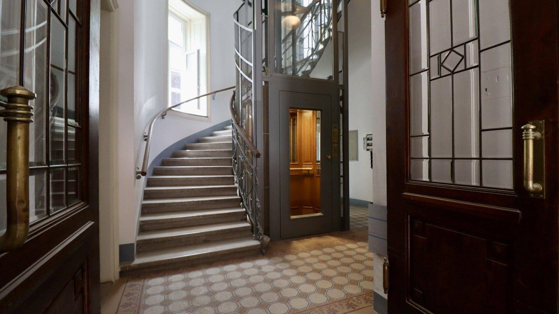 Historisches Treppenhaus mit elegantem Aufzug und gemustertem Fliesenboden in einem Altbau.