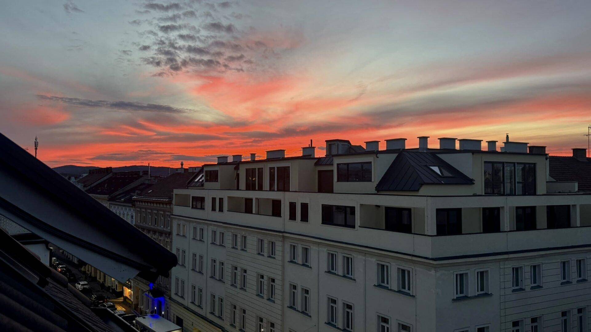 Gebäudefassade mit modernen Elementen und Dächern unter einem dramatischen Sonnenuntergangshimmel.