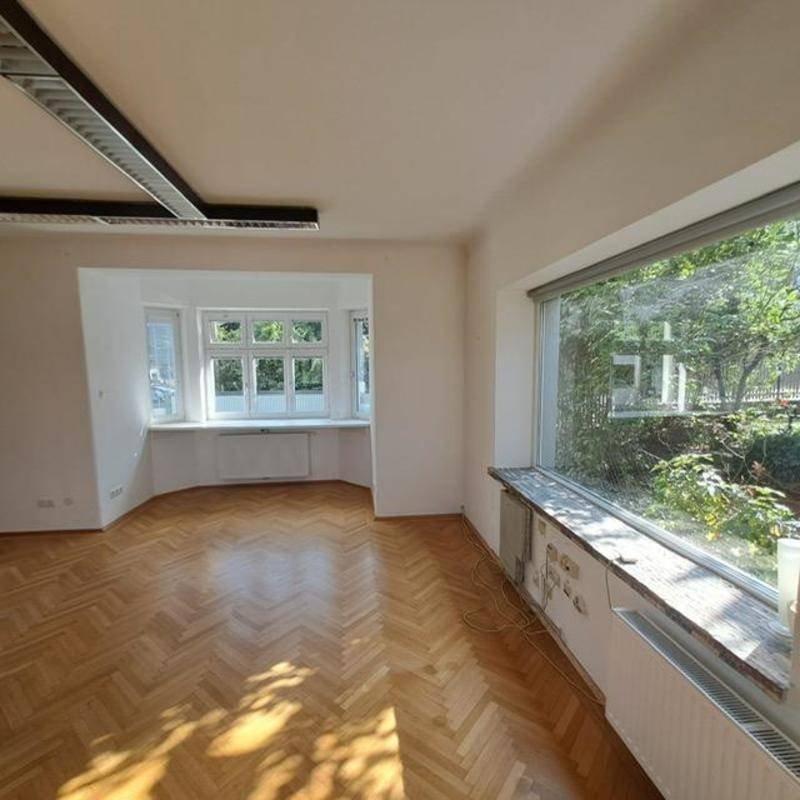 Geräumiger, heller Raum mit Fischgrätparkett und großen Fenstern, die viel Tageslicht hereinlassen.