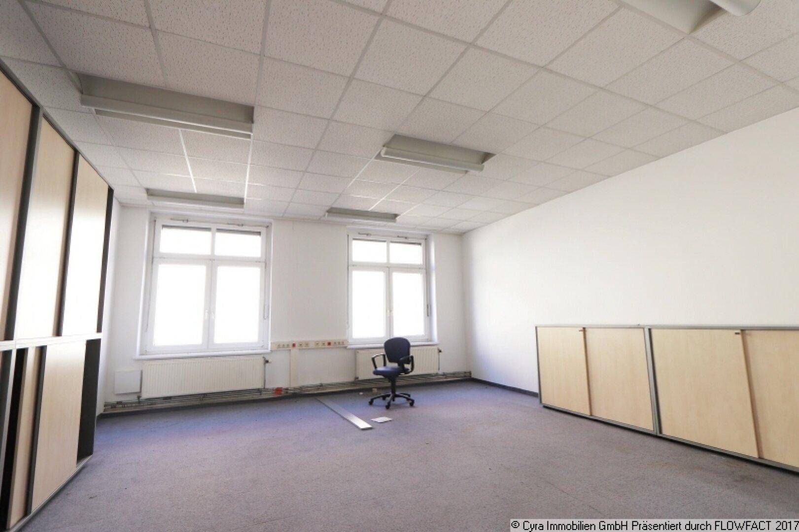 Kleineres Büro mit zwei Fenstern, Teppichboden und eingebauten Schränken an der Wand, hell und funktional.