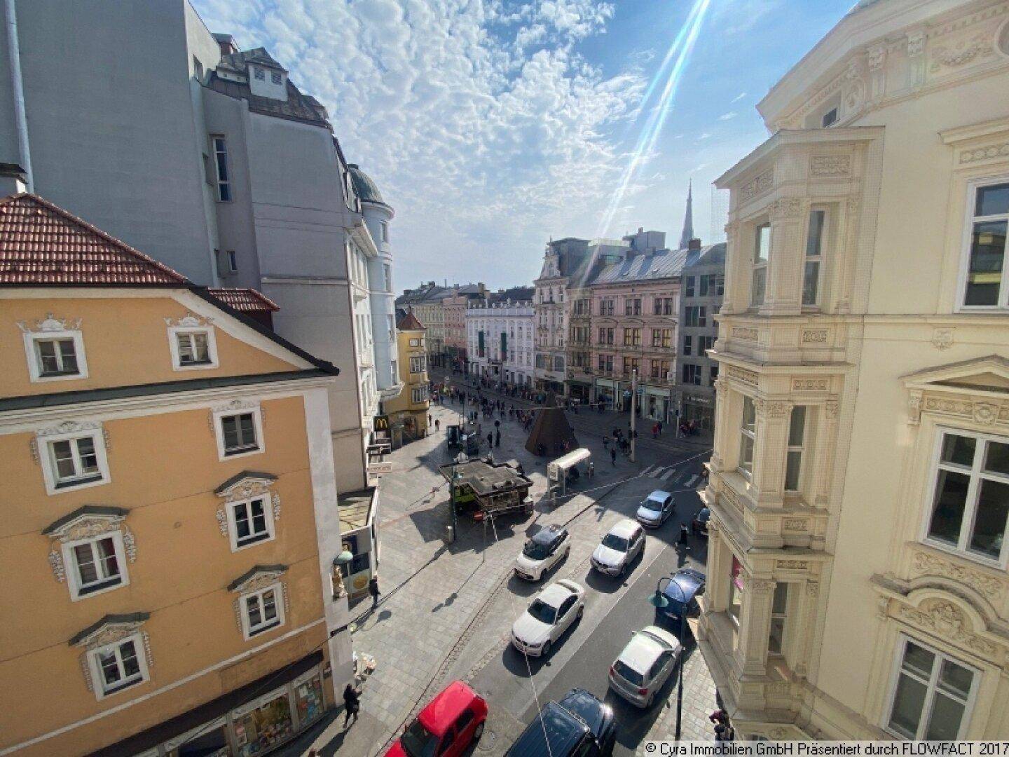Blick von oben auf eine belebte Stadtstraße mit historischen Gebäuden und parkenden Autos.