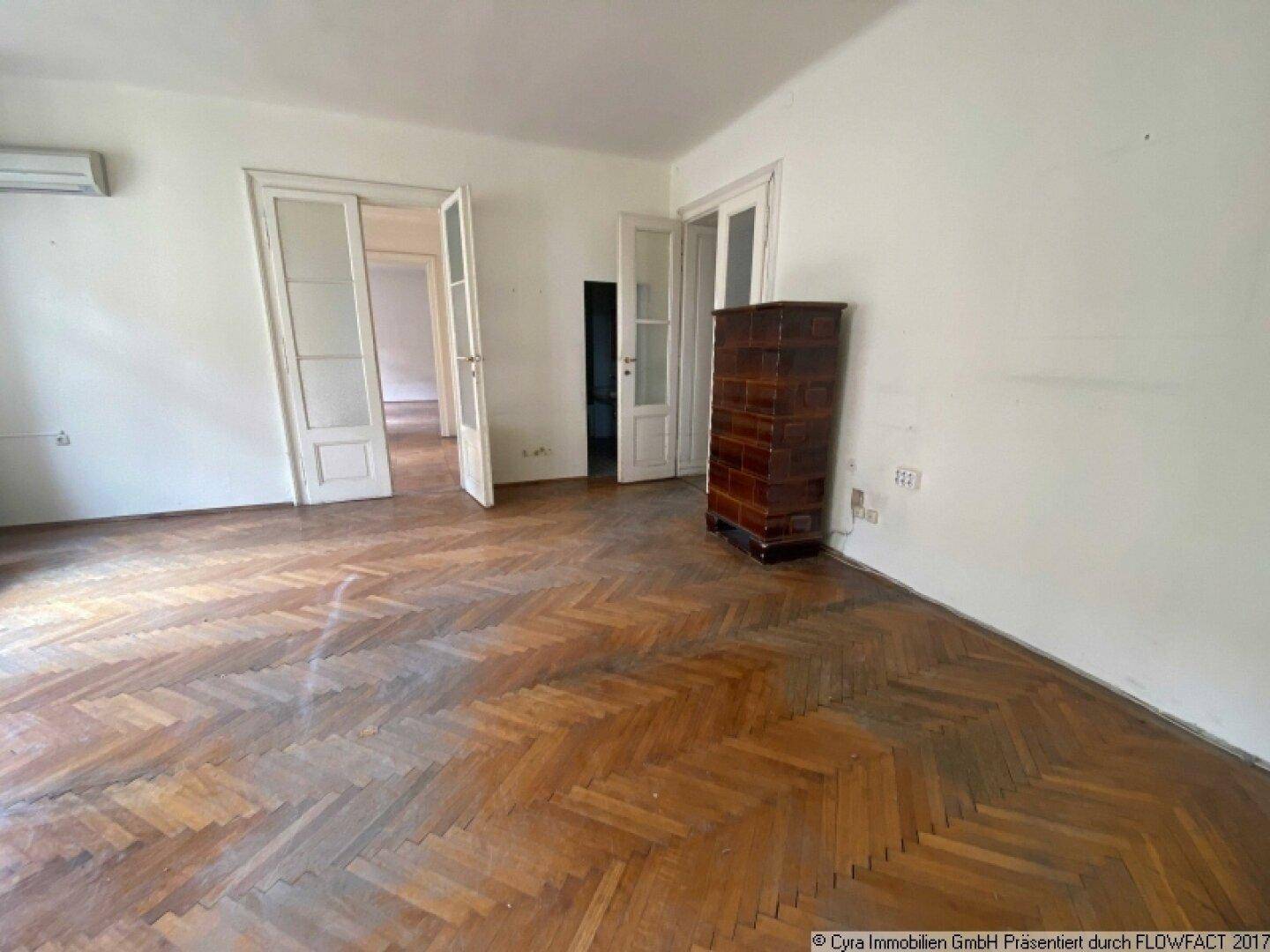 Großes Zimmer mit abgenutztem Parkettboden, hohen Decken und einem traditionellen Kachelofen.