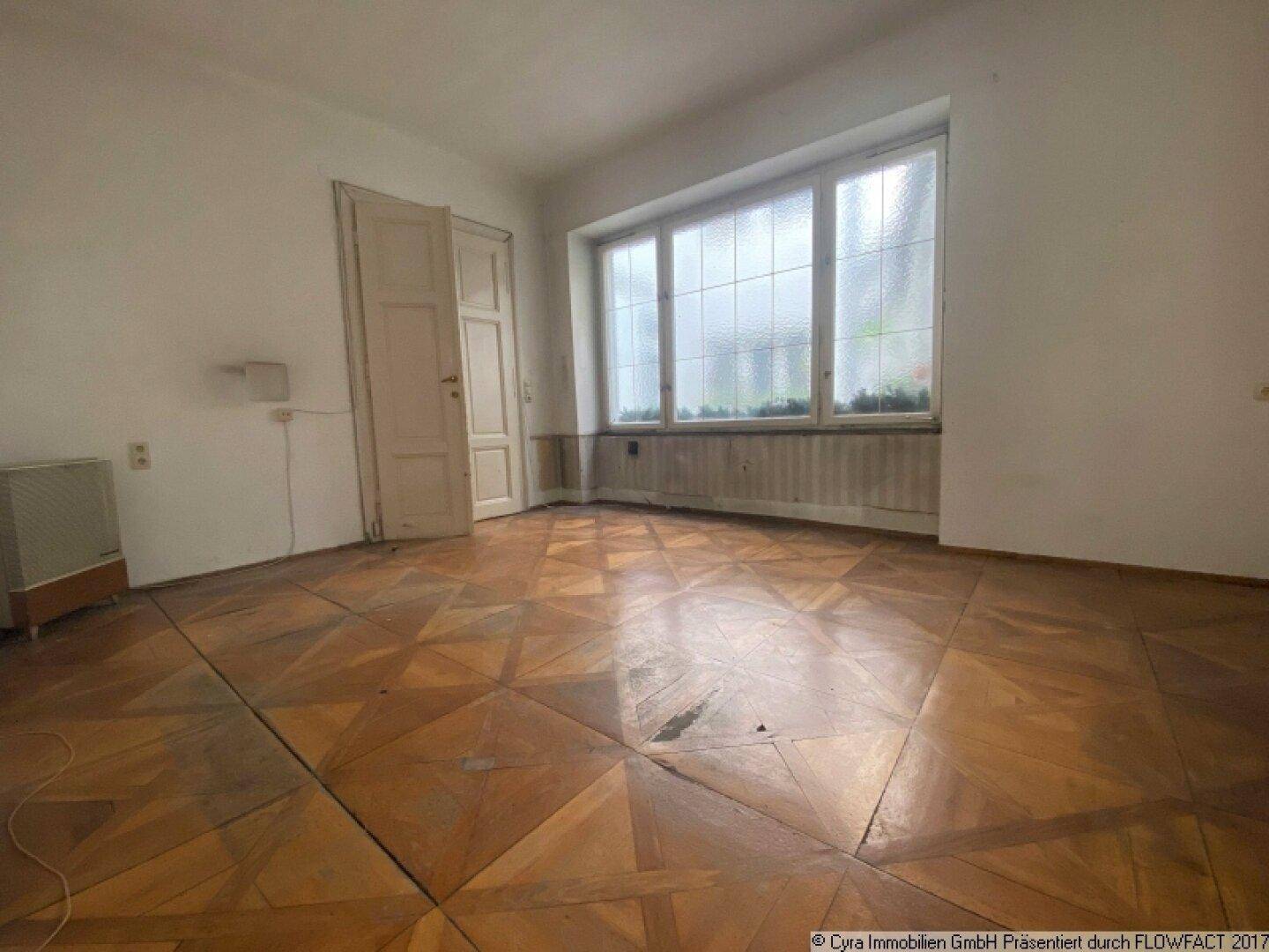 Helles Zimmer mit abgenutztem Parkettboden, großen Fenstern und einer Doppeltür, die zu einem anderen Raum führt.