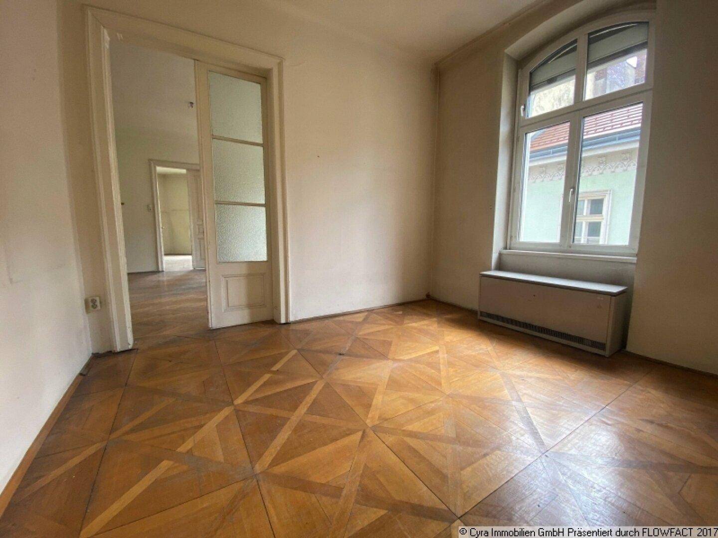 Geräumiges Zimmer mit Fischgrätparkettboden, hohen Decken und einer Doppeltür zu einem weiteren Raum.