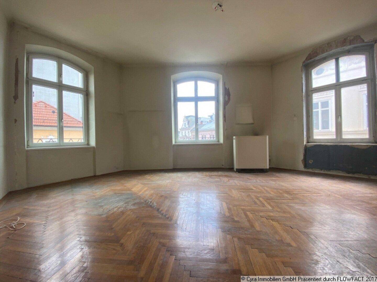 Leeres Zimmer mit abgenutztem Parkettboden, hohen Decken und drei großen Fenstern mit Stadtblick.
