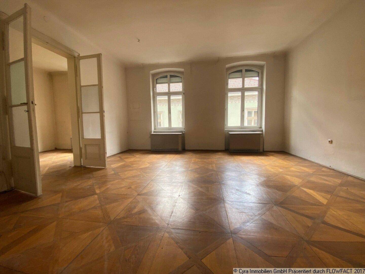 Helles Zimmer mit abgenutztem Parkettboden, hohen Decken und zwei großen Fenstern.