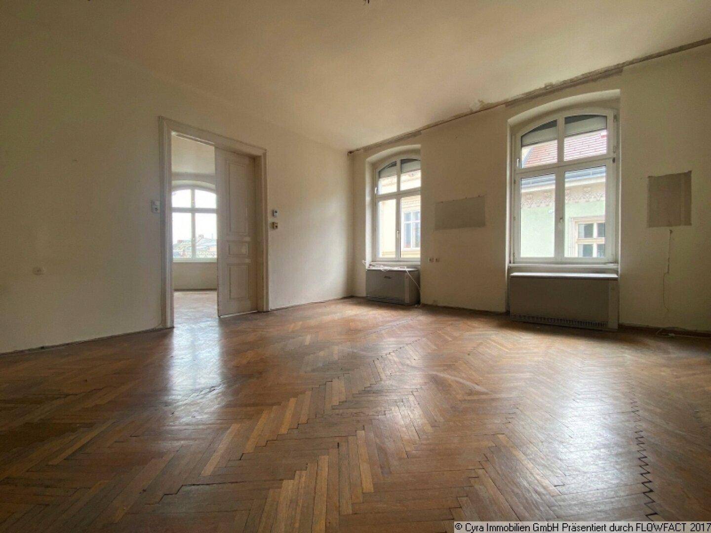 Geräumiges Zimmer mit abgenutztem Parkettboden, hohen Decken und zwei großen Fenstern.