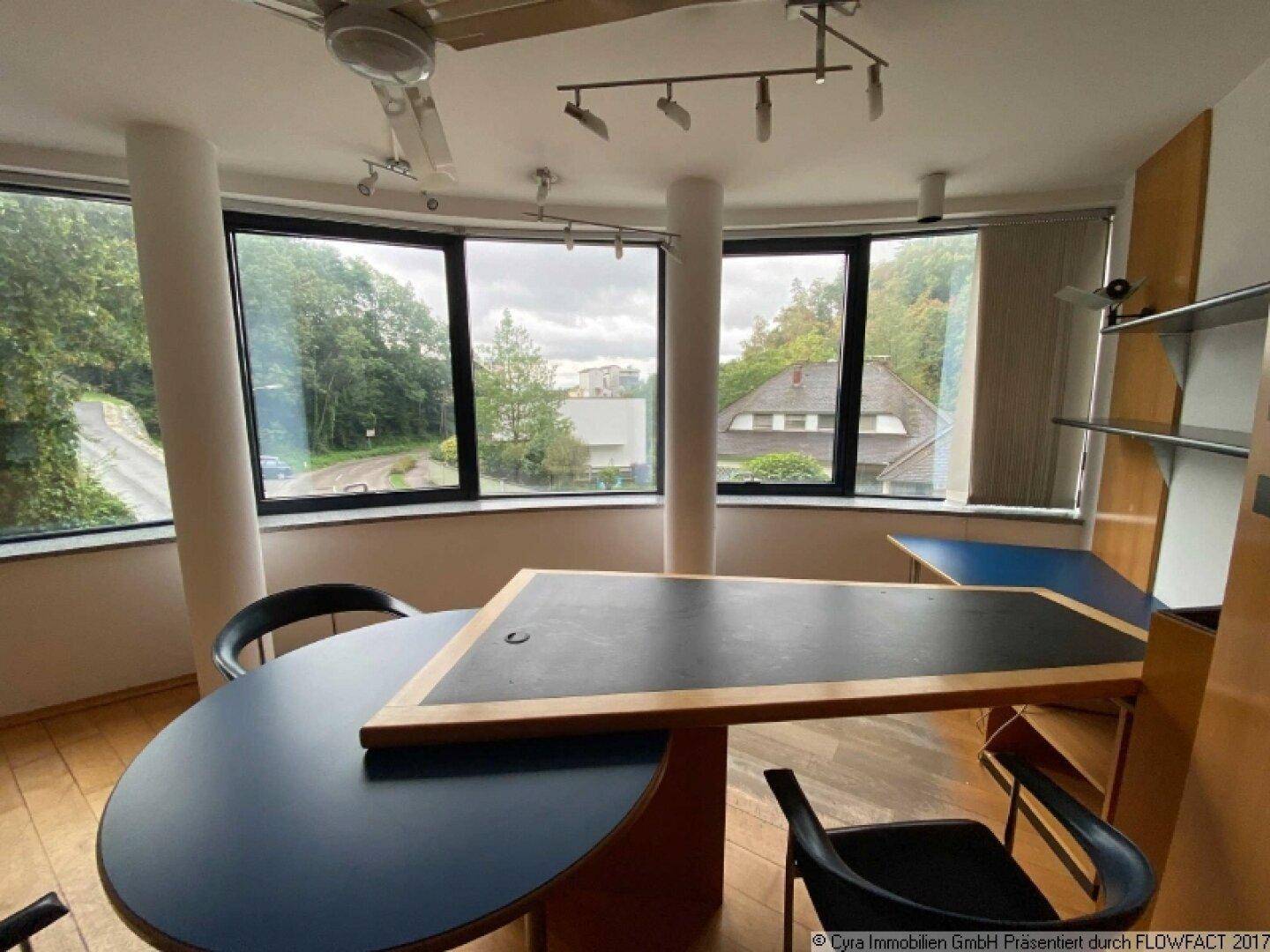 Büroraum mit mehreren Arbeitsplätzen, Parkettboden und großen Fenstern mit Blick ins Grüne.