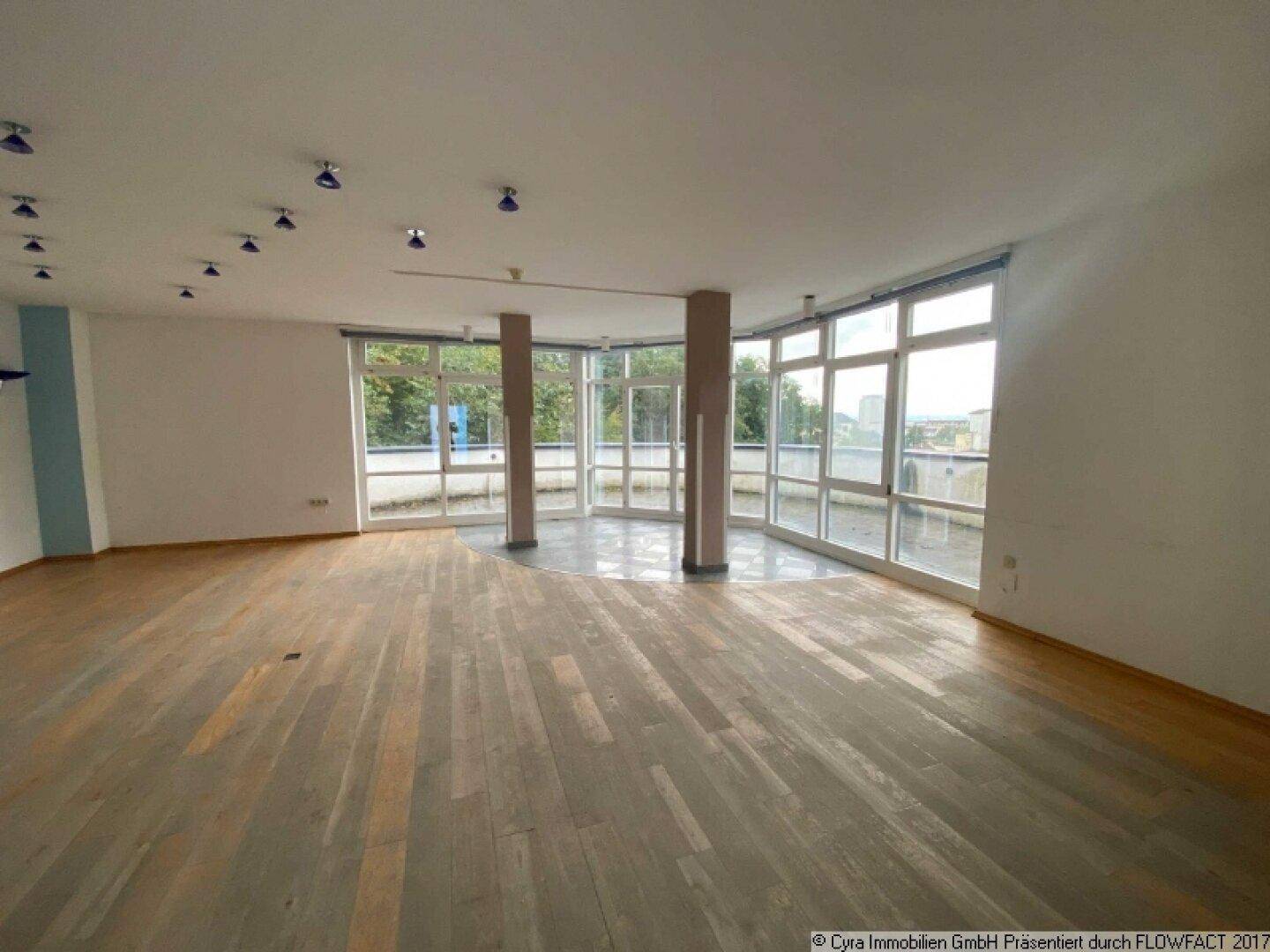 Großer, offener Raum mit Holzboden und einer umlaufenden Fensterfront, die viel Licht spendet.