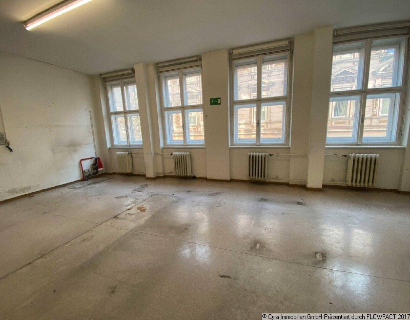 Geräumiger Raum mit mehreren großen Fenstern, Heizkörpern und einem abgenutzten Bodenbelag.