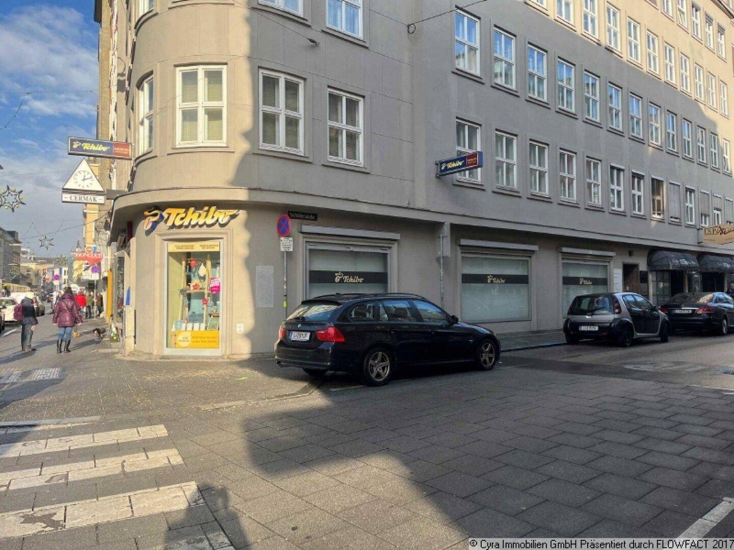 Eckgebäude mit Geschäften im Erdgeschoss, geparkten Autos und Passanten auf dem Bürgersteig.