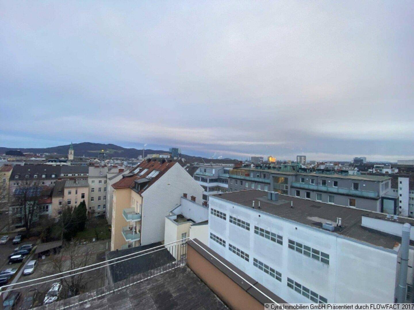 Weitblick über die Dächer der Stadt mit Wohngebäuden und Hügeln im Hintergrund unter bewölktem Himmel.