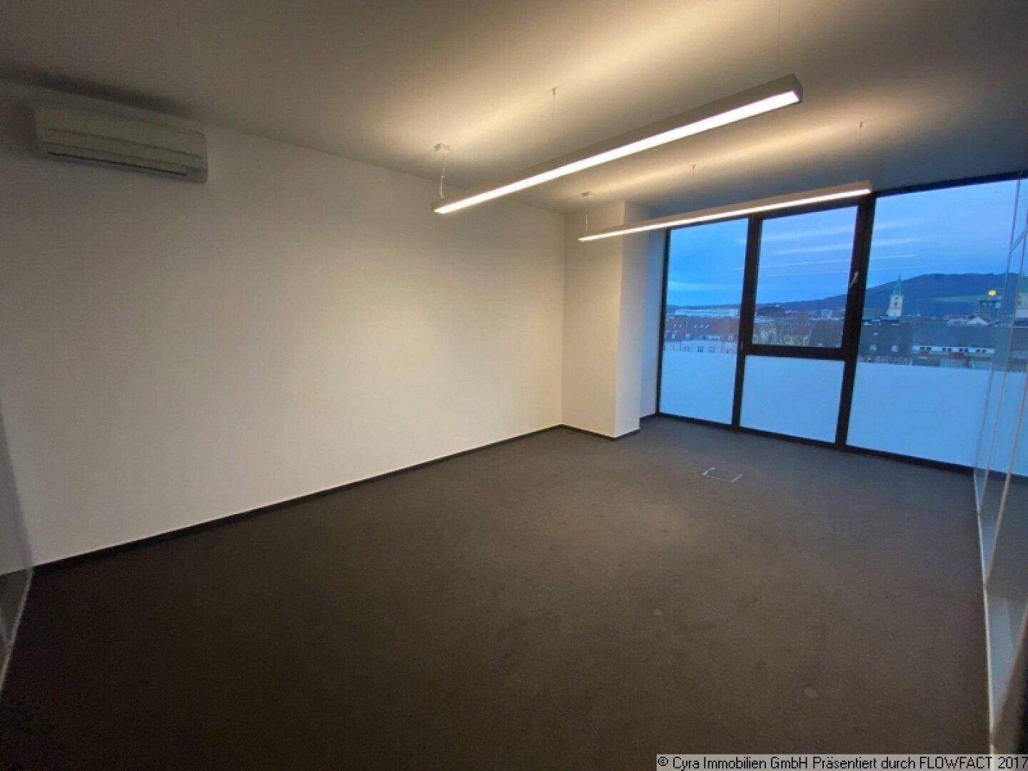 Geräumiger Büroraum mit Teppichboden, Klimaanlage und großen Fenstern mit Stadtblick.