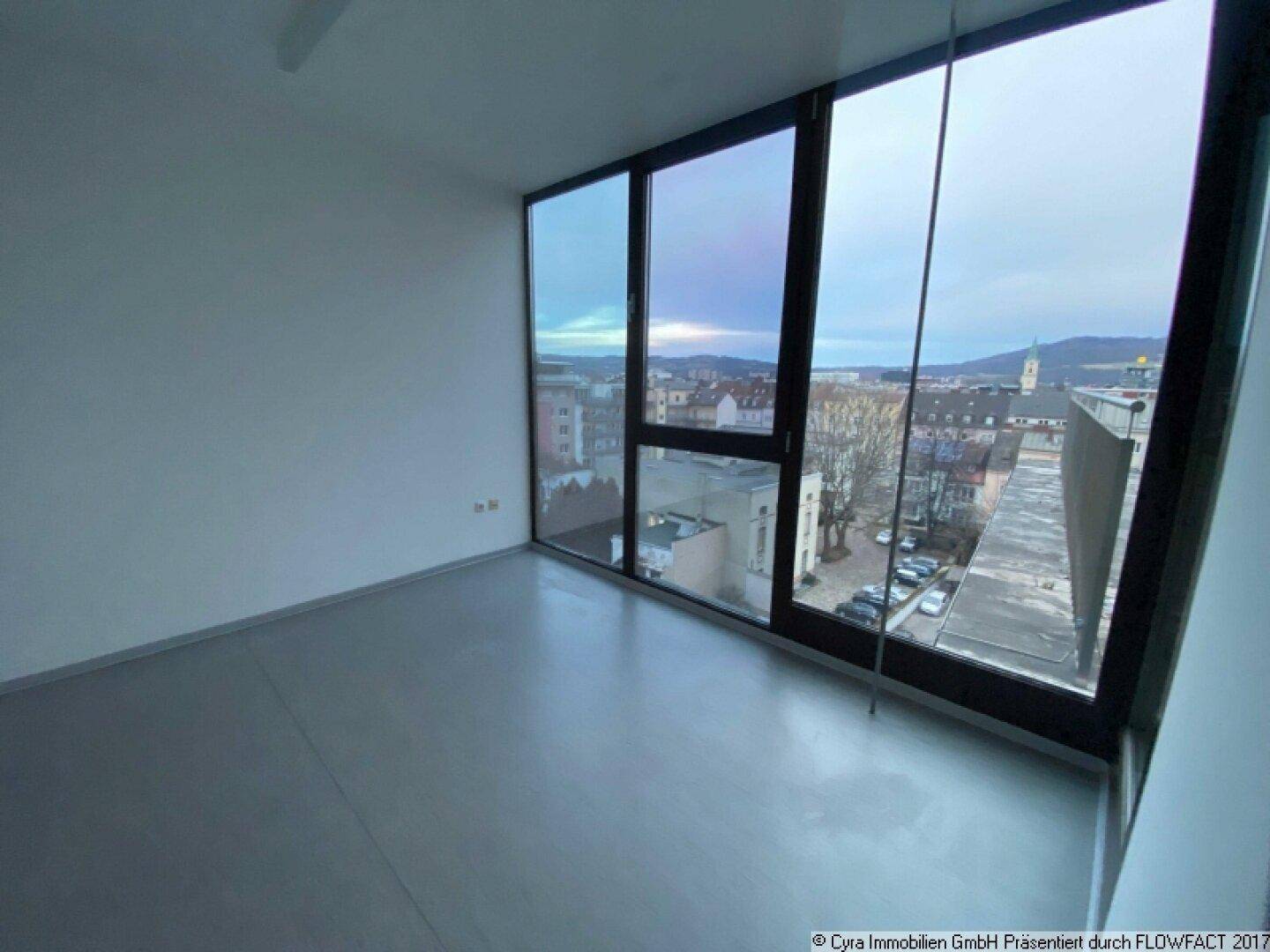 Heller Büroraum mit großen Fenstern und Blick auf die Stadt, ausgestattet mit grauem Bodenbelag.