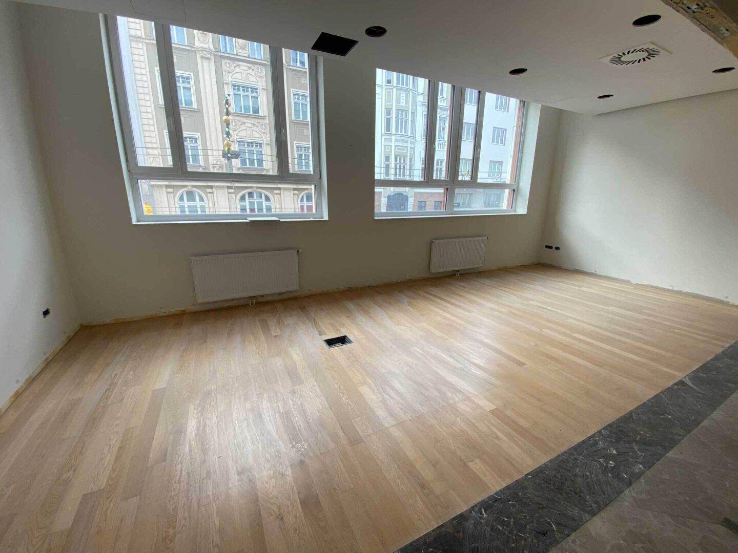 Großer Raum mit Holzparkett, Heizkörpern und Blick auf die Stadt durch hohe Fenster.