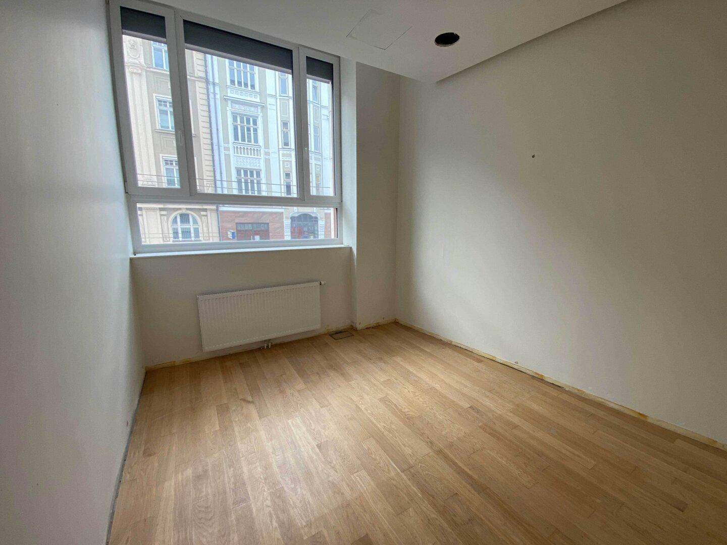 Kleinerer Raum mit Holzparkett, Heizkörper und großem Fenster mit Stadtblick, ideal für ein Büro.