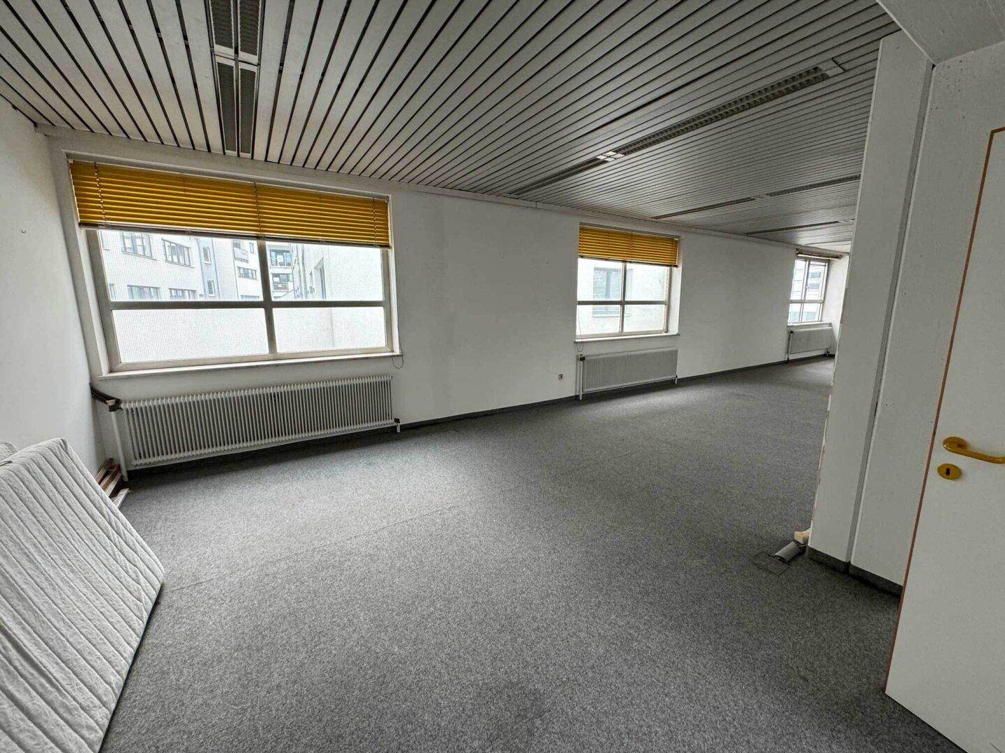 Büroraum mit grauem Teppichboden und Fenstern, die mit gelben Jalousien ausgestattet sind.