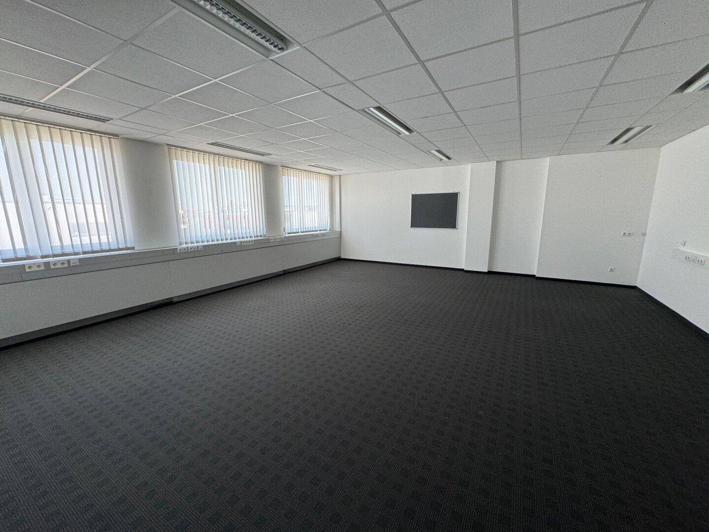 Geräumiger Büroraum mit dunklem Teppichboden, Fensterfront mit Jalousien und einer Wandtafel.